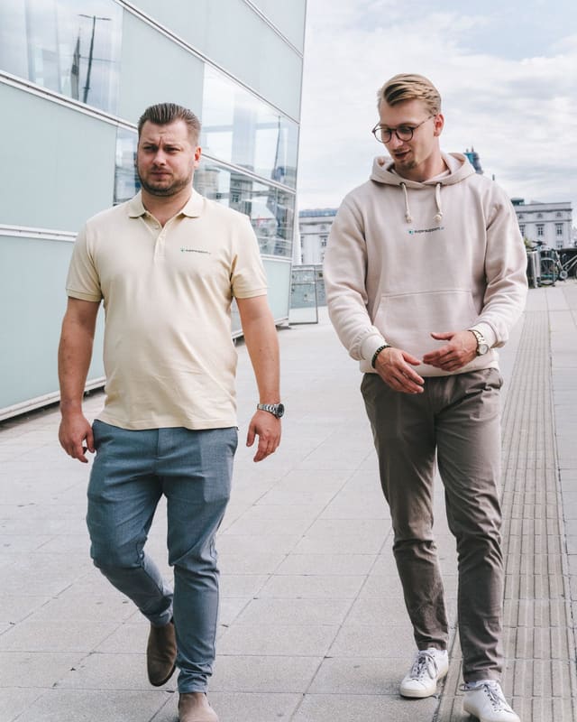 Nico und Timo Haslberger gehen im Gespräch nebeneinander an einer Glasfassade in Linz entlang